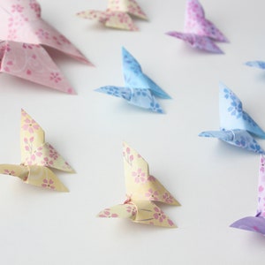May include: A collection of origami butterflies in various colors, including pink, blue, yellow, and purple. The butterflies are made of paper with a floral pattern and are arranged on a white surface.