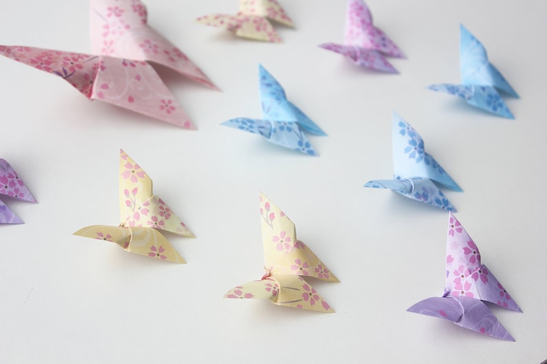 May include: A collection of origami butterflies in various colors, including pink, blue, yellow, and purple. The butterflies are made of paper with a floral pattern and are arranged on a white surface.