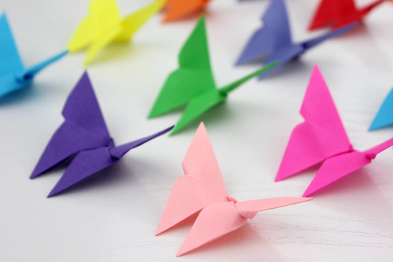 May include: A collection of colourful origami butterflies in various shades of blue, green, pink, purple, yellow and red. The butterflies are arranged on a white surface.