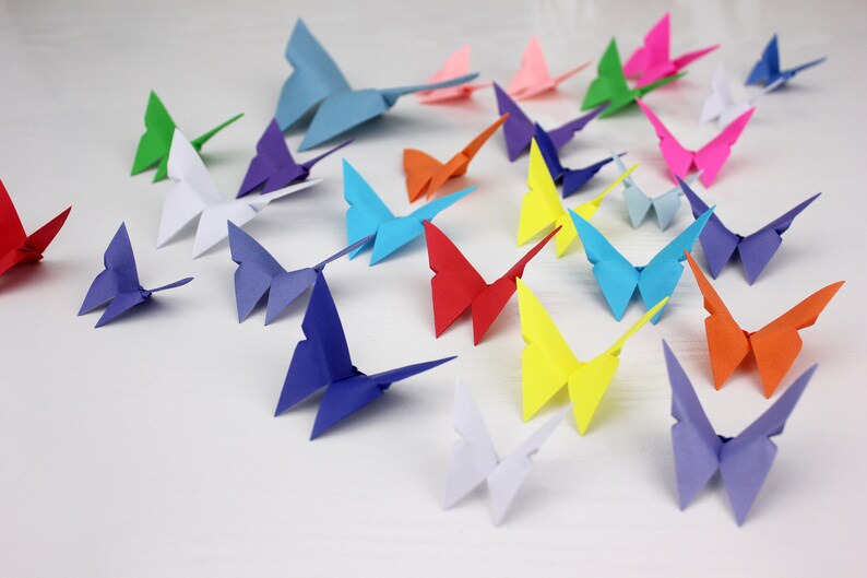 May include: A collection of colourful origami butterflies in various shades of blue, red, green, yellow, pink, and white. The butterflies are arranged on a white surface.