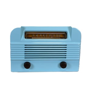 May include: A light blue radio with a horizontal speaker grille and a rectangular amber-coloured tuning dial at the top. The device features two dark brown circular control knobs positioned on the lower front panel for manual operation.