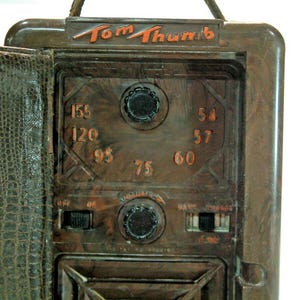 May include: A vintage brown Tom Thumb radio with a faux leather case. The radio face displays dials, frequency numbers, and the text "Volume" and "Charge". The case is open, showing the radio's interior.