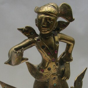 May include: A gold-coloured metal figurine of a person in a traditional dance pose, adorned with red and green gemstones. The figurine is standing on a base with a bird-like design.