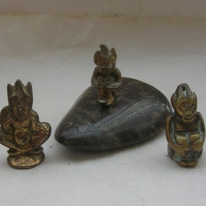 Può includere: Tre piccole statuette antiche in ottone. Una figura è seduta con una corona, un'altra è seduta su una pietra scura e la terza è in piedi. Le statuette hanno un aspetto invecchiato e sono probabilmente oggetti da collezione vintage.