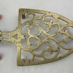 Może przedstawiać: Mosiężny podgrzewacz do naczyń z ozdobnym, ażurowym wzorem. Podgrzewacz ma uchwyt z otworem w kształcie dziurki od klucza.