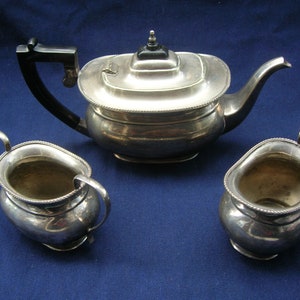 May include: A silver teapot, cream jug, and sugar bowl set. The teapot has a black handle and a silver lid. The cream jug and sugar bowl are both oval shaped.