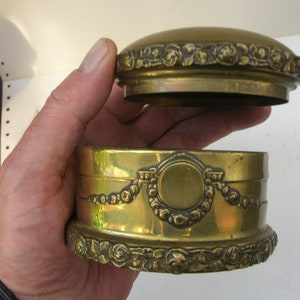 May include: A round, brass trinket box with an ornate design. The box has a hinged lid with a raised, circular design in the center. The box is decorated with a floral pattern around the edges.