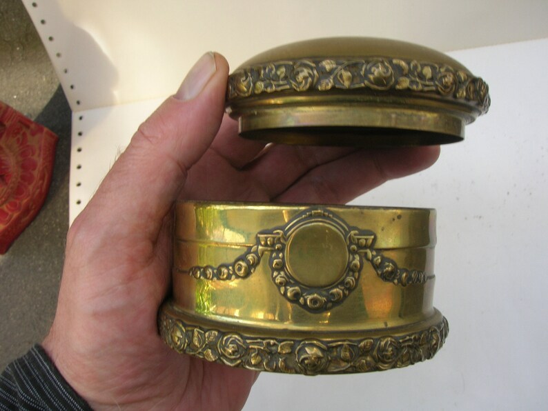 May include: A round, brass trinket box with an ornate design. The box has a hinged lid with a raised, circular design in the center. The box is decorated with a floral pattern around the edges.