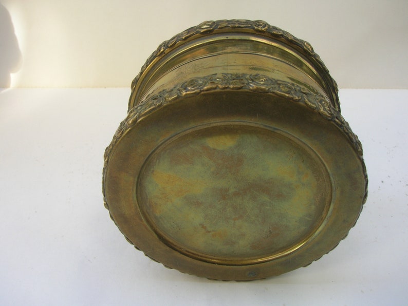 May include: A round, brass planter with a decorative, floral design around the rim. The planter has a smooth, brass bottom.