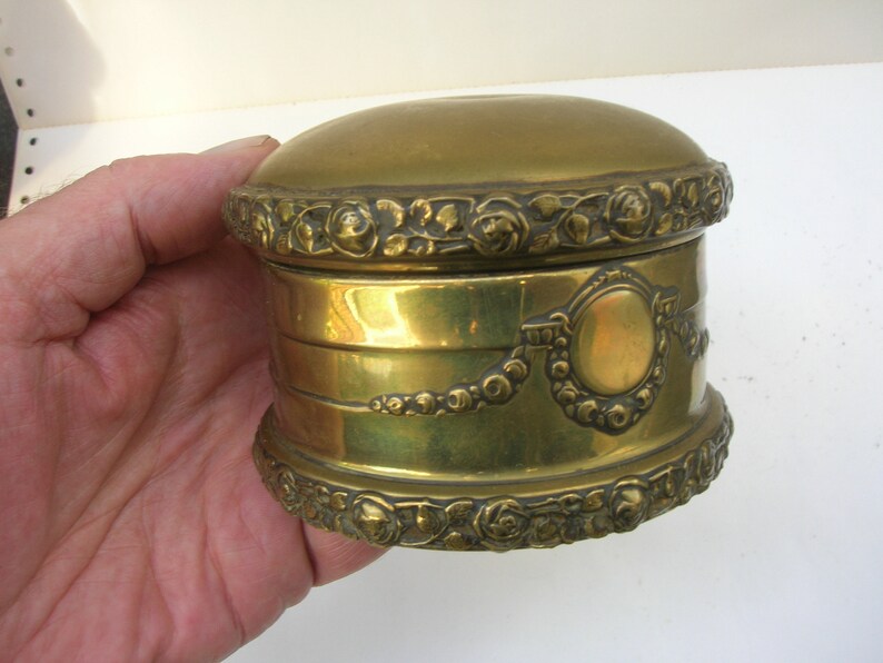 May include: A small, round, gold-colored trinket box with intricate floral designs around the top and bottom edges. The box has a raised, circular design in the center.