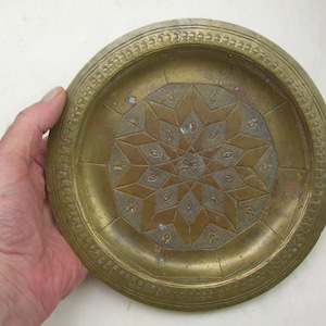 Peut inclure: Une assiette en laiton avec un motif géométrique au centre et une bordure décorative. L'assiette est circulaire et a un bord légèrement surélevé.