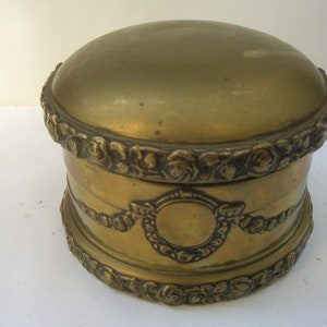 May include: A round, gold-colored metal trinket box with intricate floral and scrollwork designs around the lid and base.