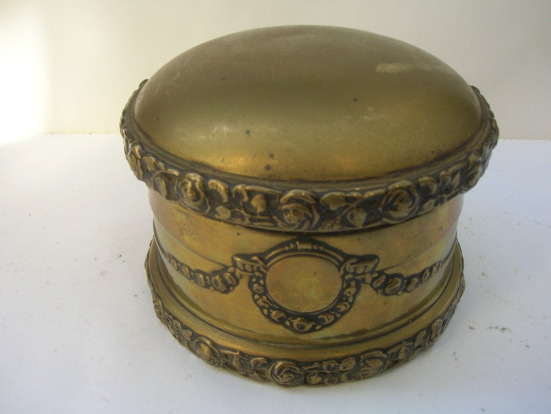 May include: A round, gold-colored metal trinket box with intricate floral and scrollwork designs around the lid and base.