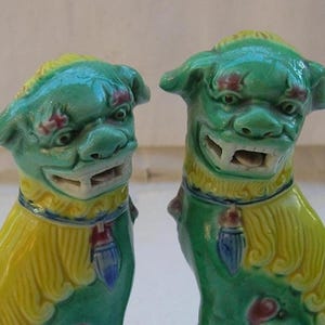Może przedstawiać: Dwie ceramiczne rzeźby psów Foo. Rzeźby są zielone z żółtymi grzywami i ogonami. Mają otwarte pyszczki i czerwone i niebieskie akcenty. Psy Foo to tradycyjna chińska figura strażnika.