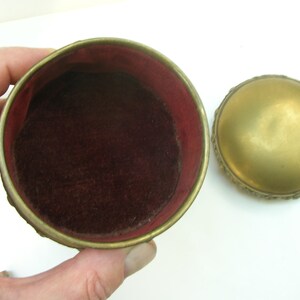 May include: A small, round, gold-colored trinket box with an ornate design. The box is open, revealing a red velvet interior.
