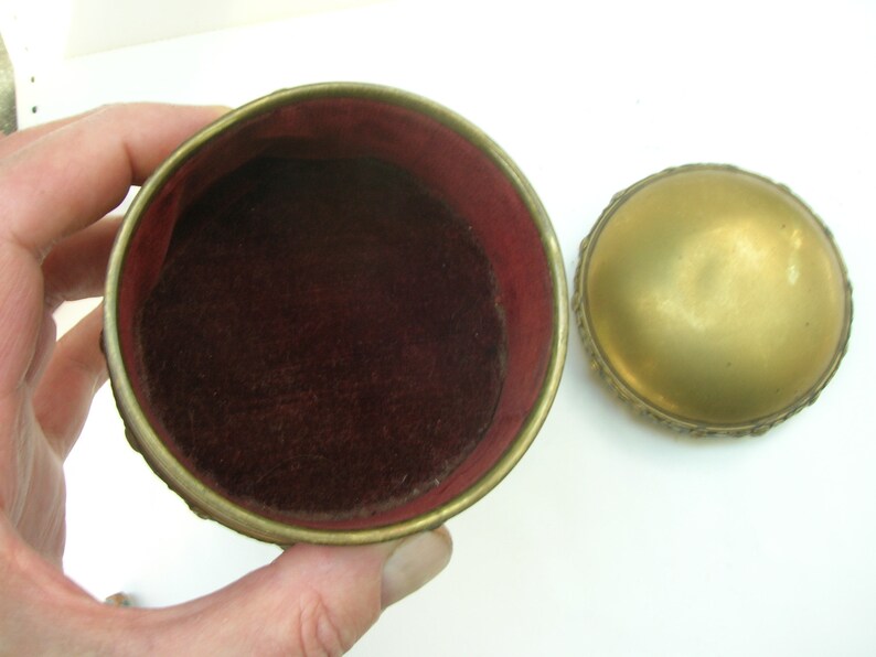 May include: A small, round, gold-colored trinket box with an ornate design. The box is open, revealing a red velvet interior.
