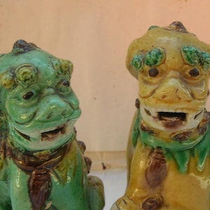 May include: Two ceramic Foo Dog statues. One is green with brown and yellow accents, the other is yellow with green and brown accents. Both have open mouths and detailed features, showcasing a traditional Asian design.