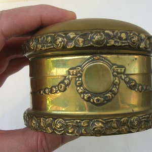 May include: A round, brass trinket box with an ornate design. The box has a hinged lid and is decorated with raised floral motifs.