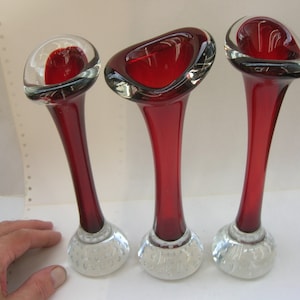 Könnte beinhalten: Drei elegante rote Glasvasen mit transparenten, blasenstrukturierten Böden. Die Vasen haben einen schlanken, gebogenen Stiel und eine ausgestellte Oberseite. Das Design ist modern und künstlerisch.