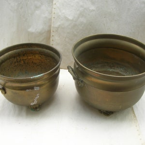 May include: Two brass planter pots with handles and feet. The larger pot is taller and wider than the smaller pot.
