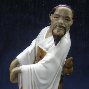 Op de afbeelding: Een keramiek beeldje van een man met een lange witte baard en een witte robe. Hij draagt een blauwe hoed en een robe met een bruin-wit patroon. Het beeldje staat en heeft een ontspannen uitdrukking.