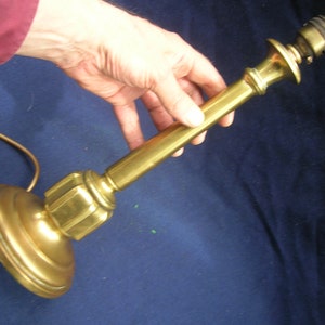 May include: A gold-toned table lamp with a simple design. The lamp has a round base and a long, slender stem.