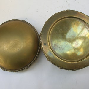 May include: A round, gold-colored metal trinket box with an ornate design around the edge. The box is open, revealing the inside of the lid and the inside of the box.