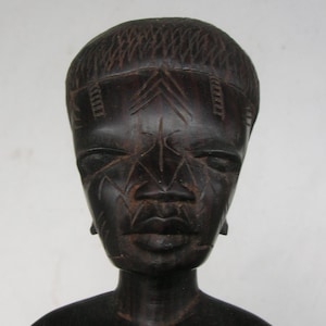 May include: A dark brown wooden sculpture of a human face and upper torso. The face has carved details including lines across the face, a defined nose, and closed eyes. The hair is textured and the sculpture has a smooth finish.