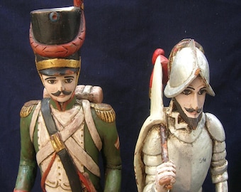 Op de afbeelding: Twee handgesneden houten beeldjes van historische figuren. De ene is een soldaat in een groen en wit uniform met een zwarte hoed. De andere is een conquistador in zilveren harnas en helm, met een staf met een rood en witte vlag.