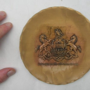 May include: A small, round, gold-coloured metal plate with a raised design of the Pennsylvania state seal. The seal features two horses, a ship, and a crest. The text "Commonwealth of Pennsylvania" is inscribed below the seal.