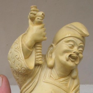 May include: A carved ivory figurine of a smiling man wearing a hat and holding a staff. The figurine is detailed with intricate carvings and has a warm, creamy color.