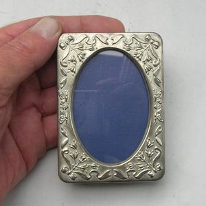 May include: A rectangular silver-toned metal picture frame with rounded corners and an oval centre opening. The frame features an embossed decorative border with floral motifs, ribbons, and small butterfly shapes surrounding a dark blue insert.