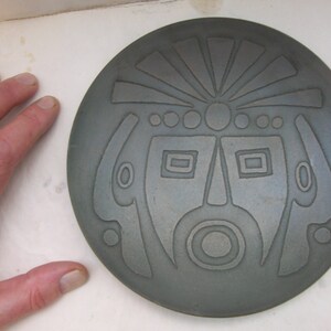May include: A round, gray ceramic plate with a stylized face design. The face has a headdress, eyes, a nose, and a mouth.