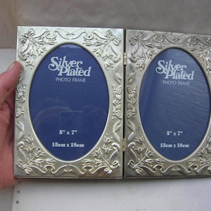 Peut inclure: Cadre photo double argenté avec ouvertures ovales et un motif floral décoratif. Chaque cadre mesure 13cm x 18cm et a un fond bleu. Le texte "Silver Plated PHOTO FRAME" est visible sur chaque cadre.