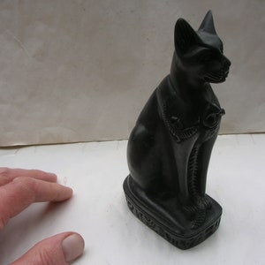 May include: Black Egyptian cat statue with intricate details. The cat is sitting with its head turned to the side. The statue is on a base with hieroglyphics.