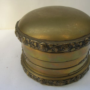 May include: A gold metal trinket box with a round lid and intricate floral designs around the base.