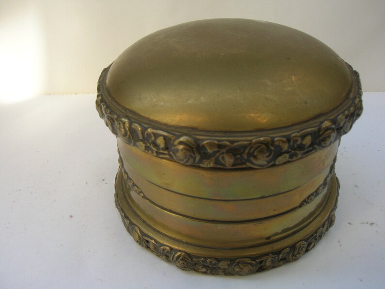 May include: A gold metal trinket box with a round lid and intricate floral designs around the base.