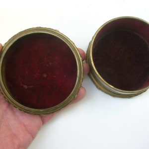 May include: Two round, gold-colored trinket boxes with red velvet interiors. The boxes are open and the lids are off.
