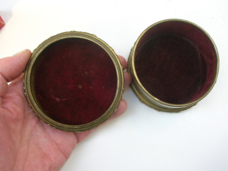 May include: Two round, gold-colored trinket boxes with red velvet interiors. The boxes are open and the lids are off.