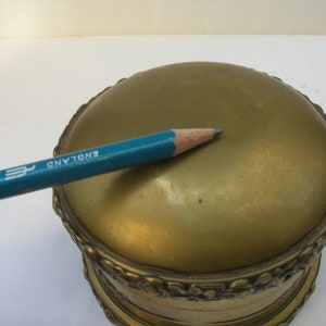 May include: A gold-colored round trinket box with an ornate design around the edge. The top of the box is smooth and has the words "ENGLAND" inscribed on it.