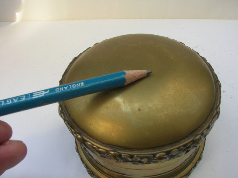May include: A gold-colored round trinket box with an ornate design around the edge. The top of the box is smooth and has the words "ENGLAND" inscribed on it.