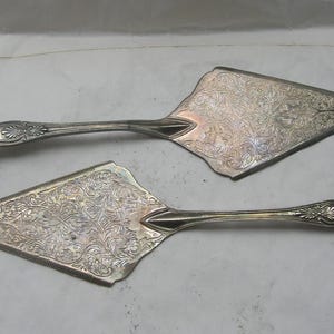 Può includere: Due palette per torta d'argento con disegni incisi decorativi. Le palette hanno una lama appuntita e un manico decorativo.