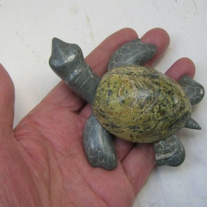 Pode incluir: Uma escultura de tartaruga de pedra esculpida à mão com uma carapaça verde e cinza manchada. A tartaruga está virada para a direita e tem uma cabeça e pernas detalhadas.