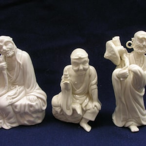 Peut inclure: Trois figurines en céramique blanc cassé représentant des personnages en robes traditionnelles. La figure de gauche est assise sur un rocher, celle du centre est assise en tenant un petit objet, et celle de droite porte un baluchon sur l'épaule.