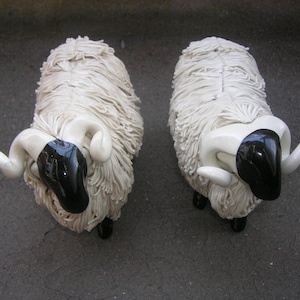 Op de afbeelding: Twee witte keramische schapenfiguren met zwarte gezichten en hoorns. De schapen staan op een grijs oppervlak.
