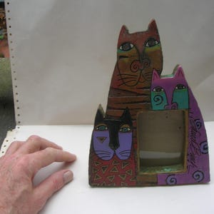 May include: A ceramic picture frame featuring three stylized cats. The cats are stacked on top of each other, with the largest cat at the bottom. The cats are painted in various colors, including purple, blue, red, and brown. The frame has a rectangular opening for a photo.