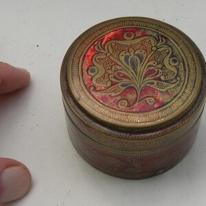 Puede incluir: Una caja redonda de latón antiguo con interior rojo y diseños florales y de aves intrincados. La tapa y los lados presentan grabados detallados, creando una estética vintage. La caja probablemente se utiliza para joyas o pequeños recuerdos.