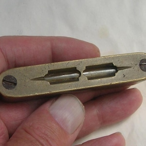 May include: A vintage brass tool, rectangular in shape, with two parallel recessed channels. Each channel has a central, pointed metal piece. Two screws secure the ends. The tool is held in a hand.
