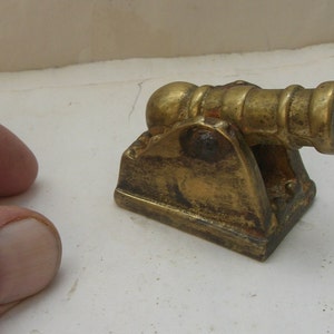 Puede incluir: Un pequeño cañón de metal dorado sobre una base. El cañón está detallado con una bola redonda al final del cañón y un diseño decorativo en la base.