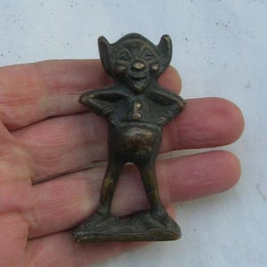 Może przedstawiać: Mała, brązowa figurka przedstawiająca gobliny z ostrymi uszami, przebiegłym uśmiechem i okrągłym brzuchem. Goblin stoi z założonymi rękami i ma na sobie prostą tunikę.